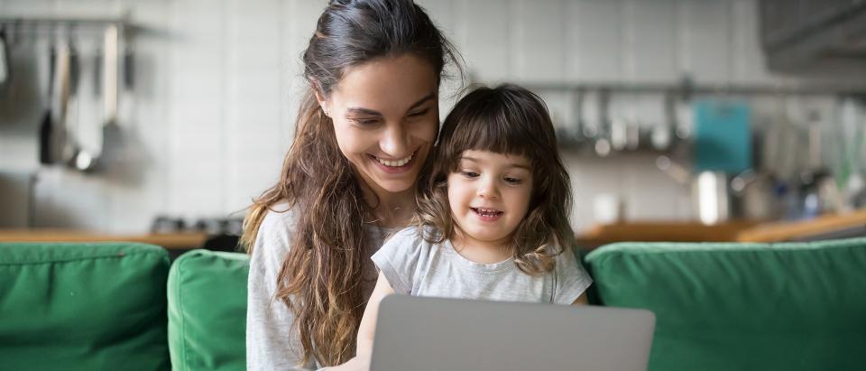 Woman sitting on a coach with a toddler girl on her lap, looking at a laptop and smiling.