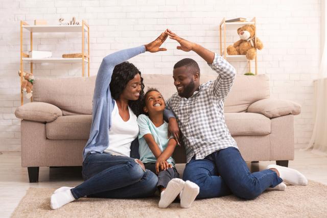 Man and woman sitting on the floor, holding their arm up in the shape of a house with child sitting in the middle.