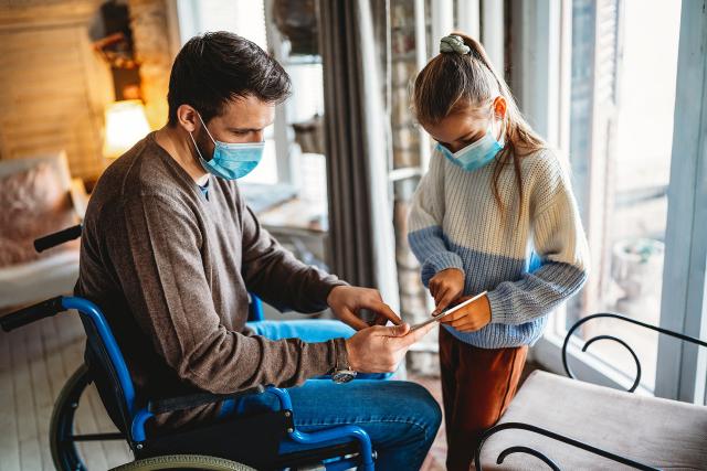 Man in wheelchair with mask showing a tablet to a girl wearing a mask