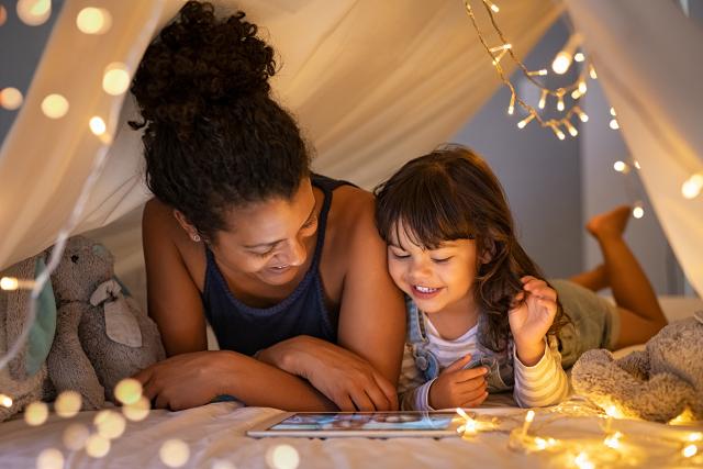Woman and girl laying on their bellies inside a tent with fairy lights, watching a tablet